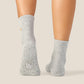 A pair of light gray ankle socks with white grip dots on the soles, shown worn on a person's feet against a solid beige background.
