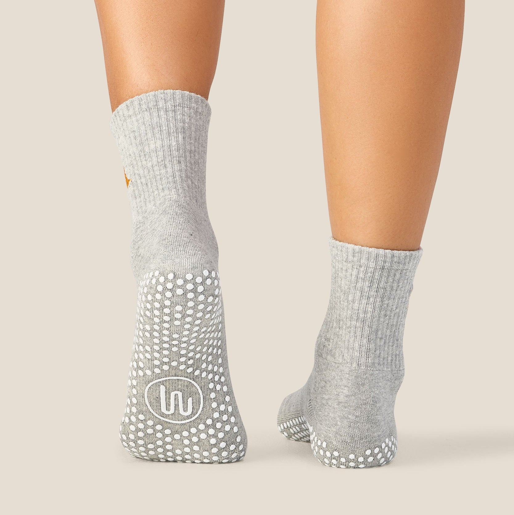 A pair of light gray ankle socks with white grip dots on the soles, shown worn on a person's feet against a solid beige background.